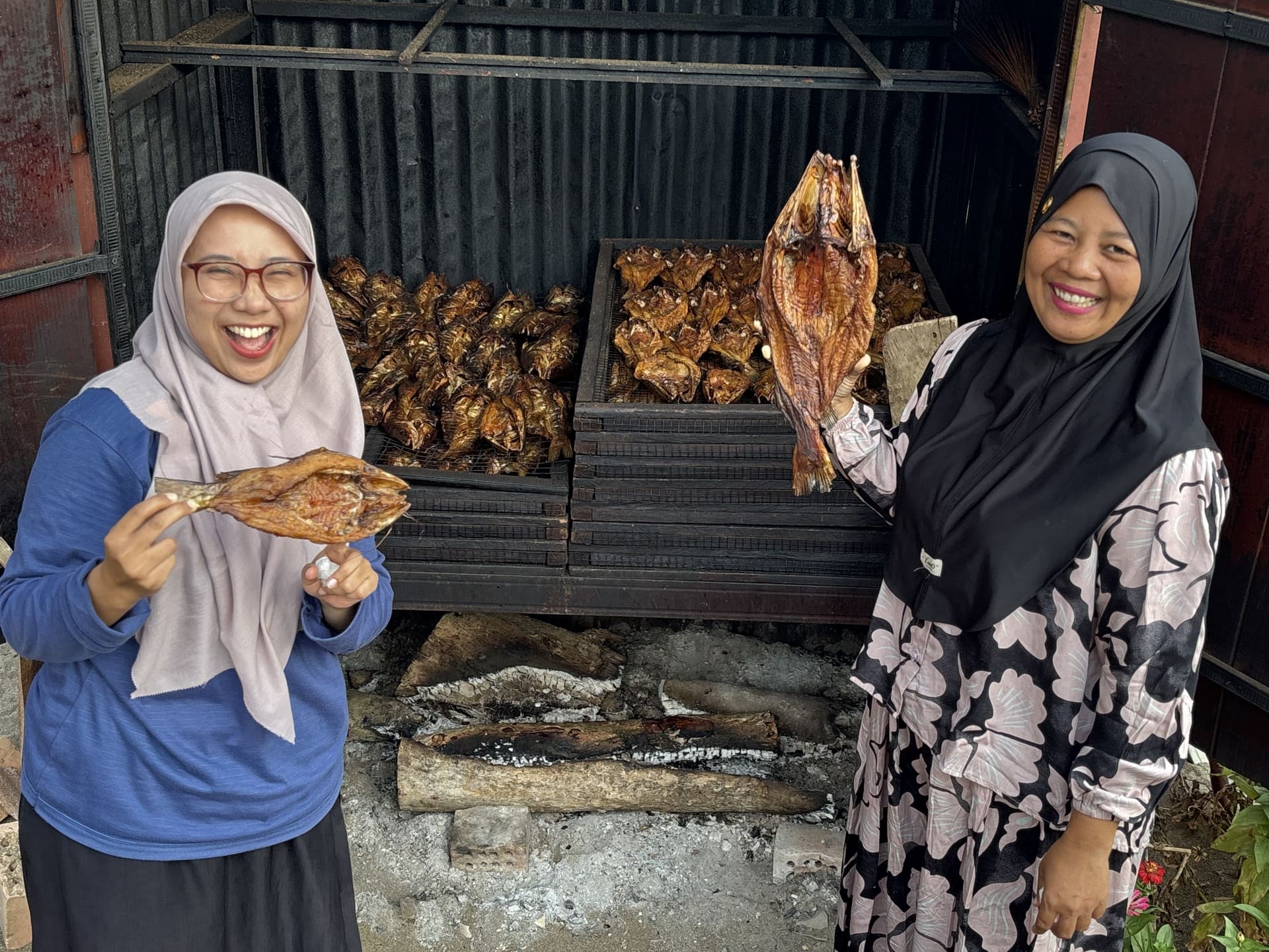 Two women smiling as they hold up freshly smoked fish in front of a smokehouse